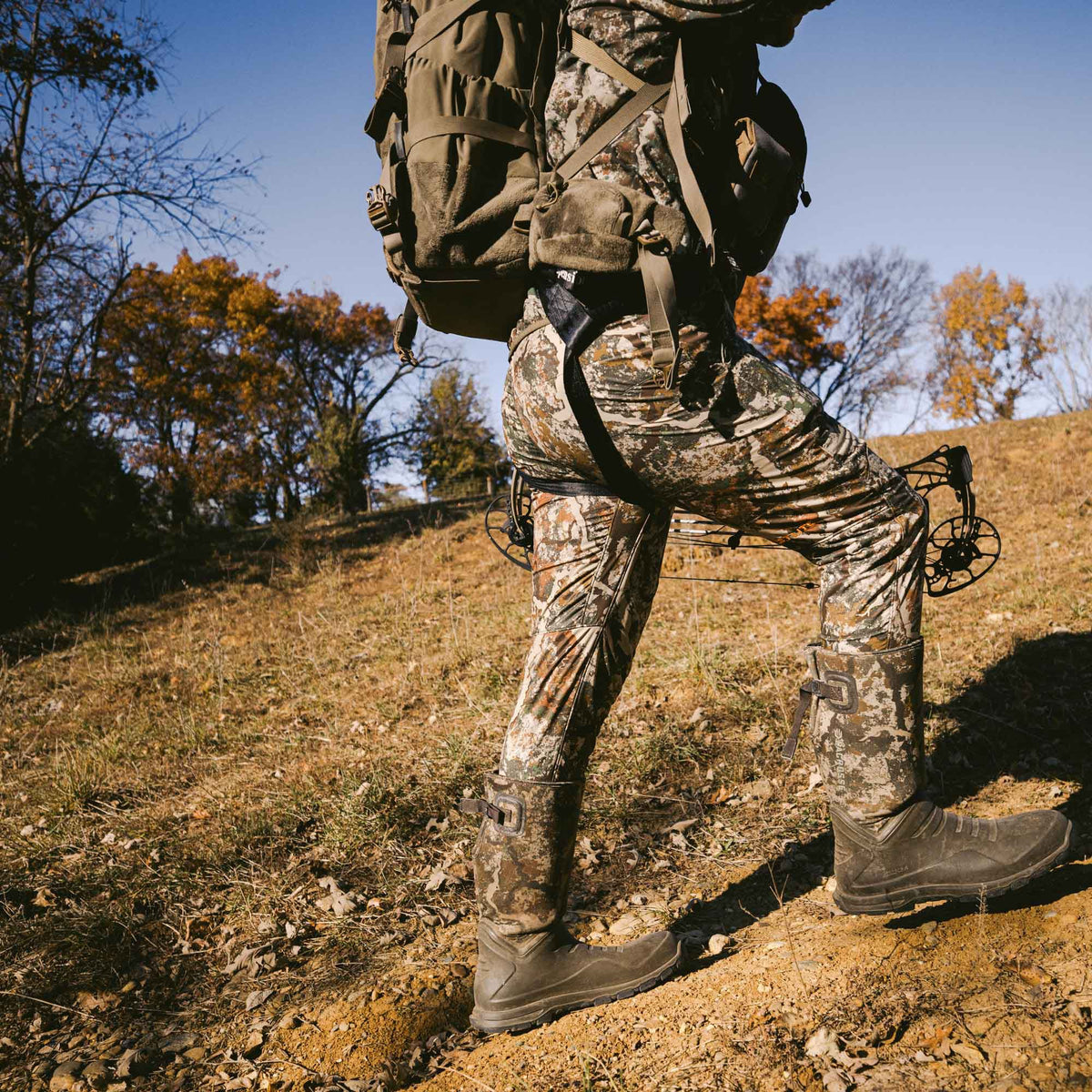 Person in camouflage hunting pants and dark rubber boots with camo shafts carries a brown backpack and bow uphill.