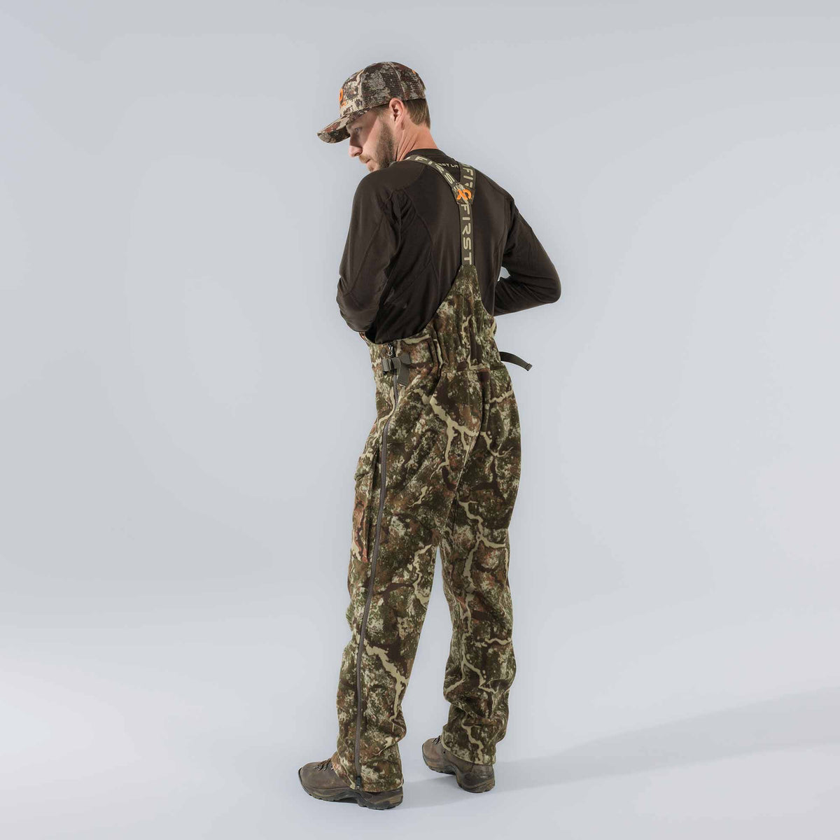 Brown and green camouflage hunting bibs with full-length side zippers and First Lite branded suspenders.