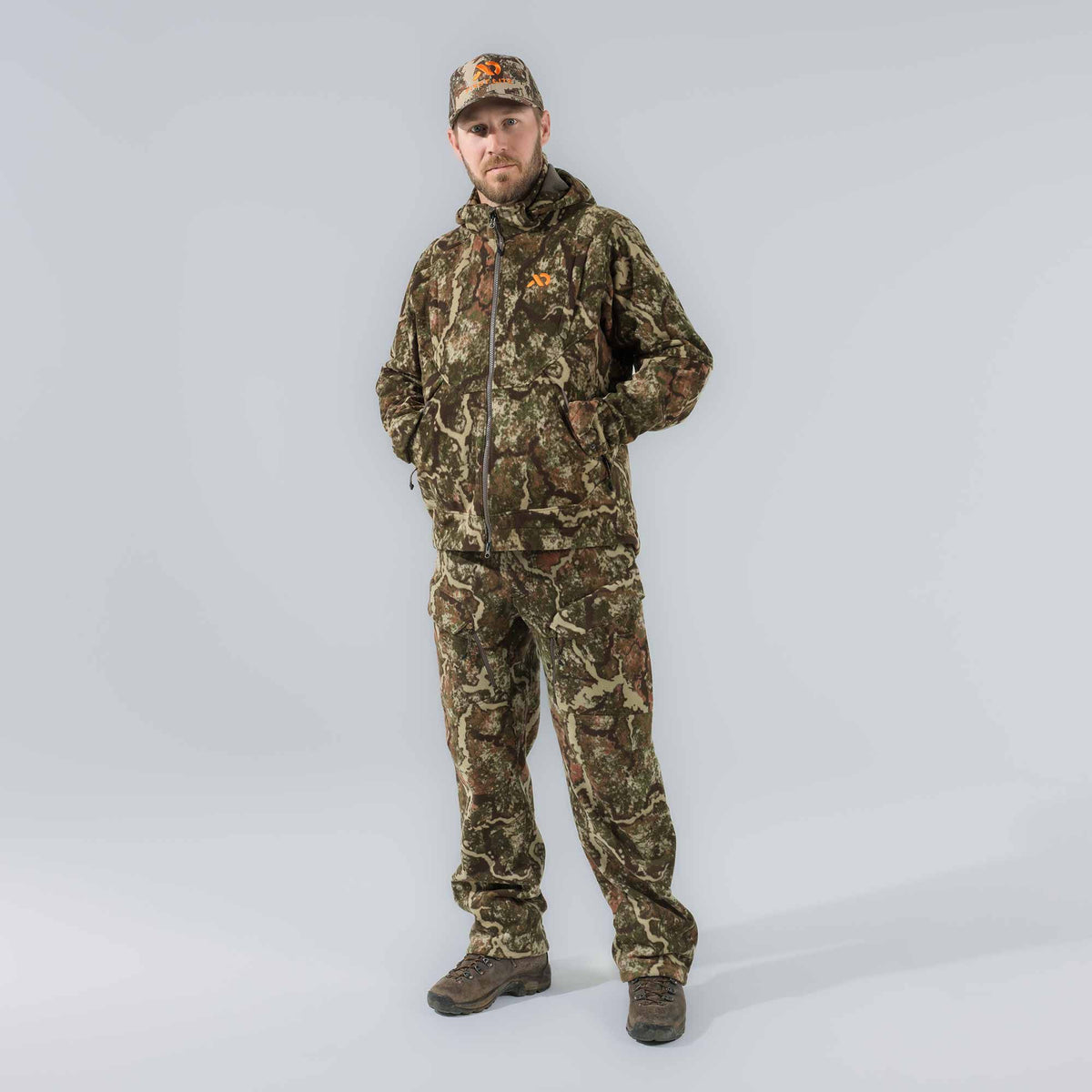 A man wears a camouflage hunting suit, featuring a hooded full-zip jacket, matching pants with cargo pockets, and a cap with an orange logo.