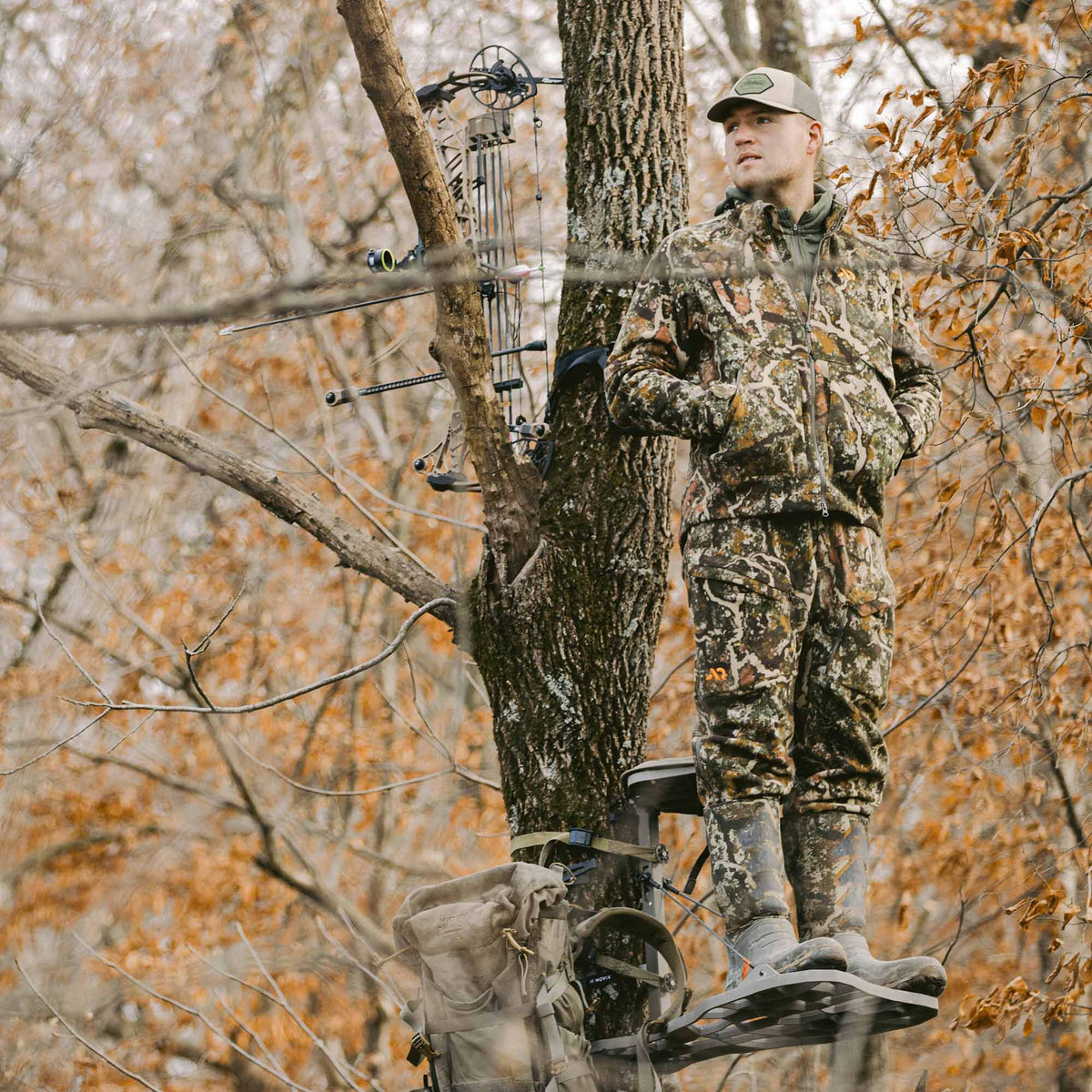 A hunter in full multi-color camouflage gear stands on a grey metal tree stand, with a bow and tan backpack visible on the tree.