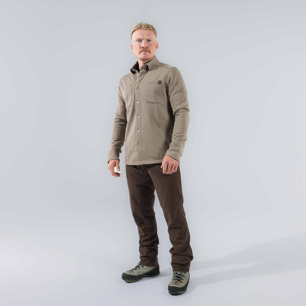 A men's taupe long-sleeve collared shirt with snap buttons, two chest pockets, and a black logo patch on the right chest pocket.
