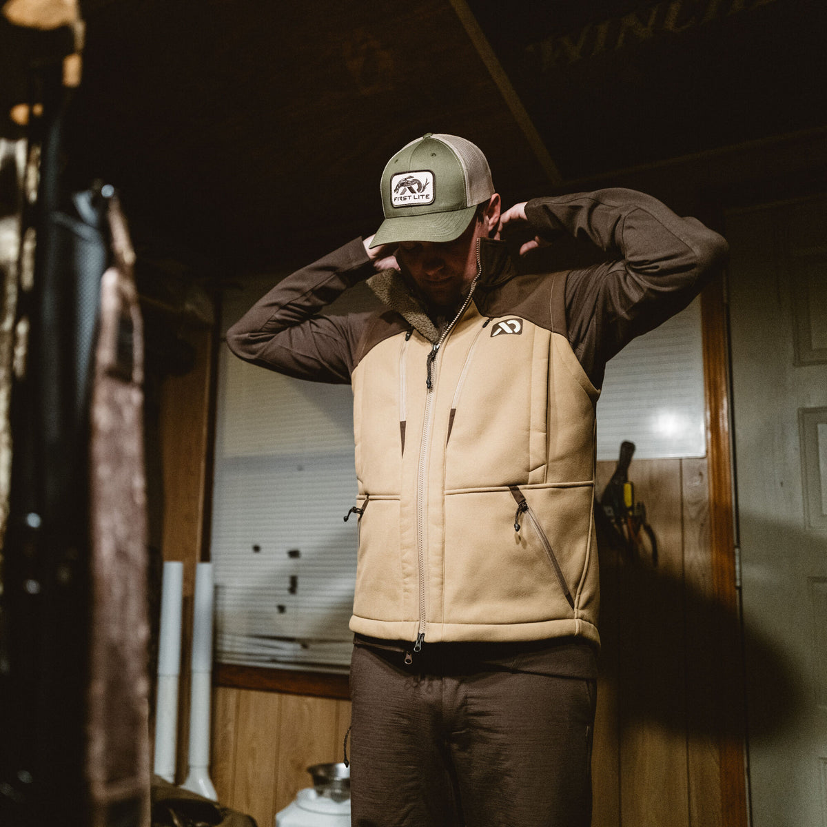 First Lite two-tone tan and brown insulated vest featuring a full-zip front, zippered pockets, a fleece-lined collar, and an olive green logoed mesh cap.
