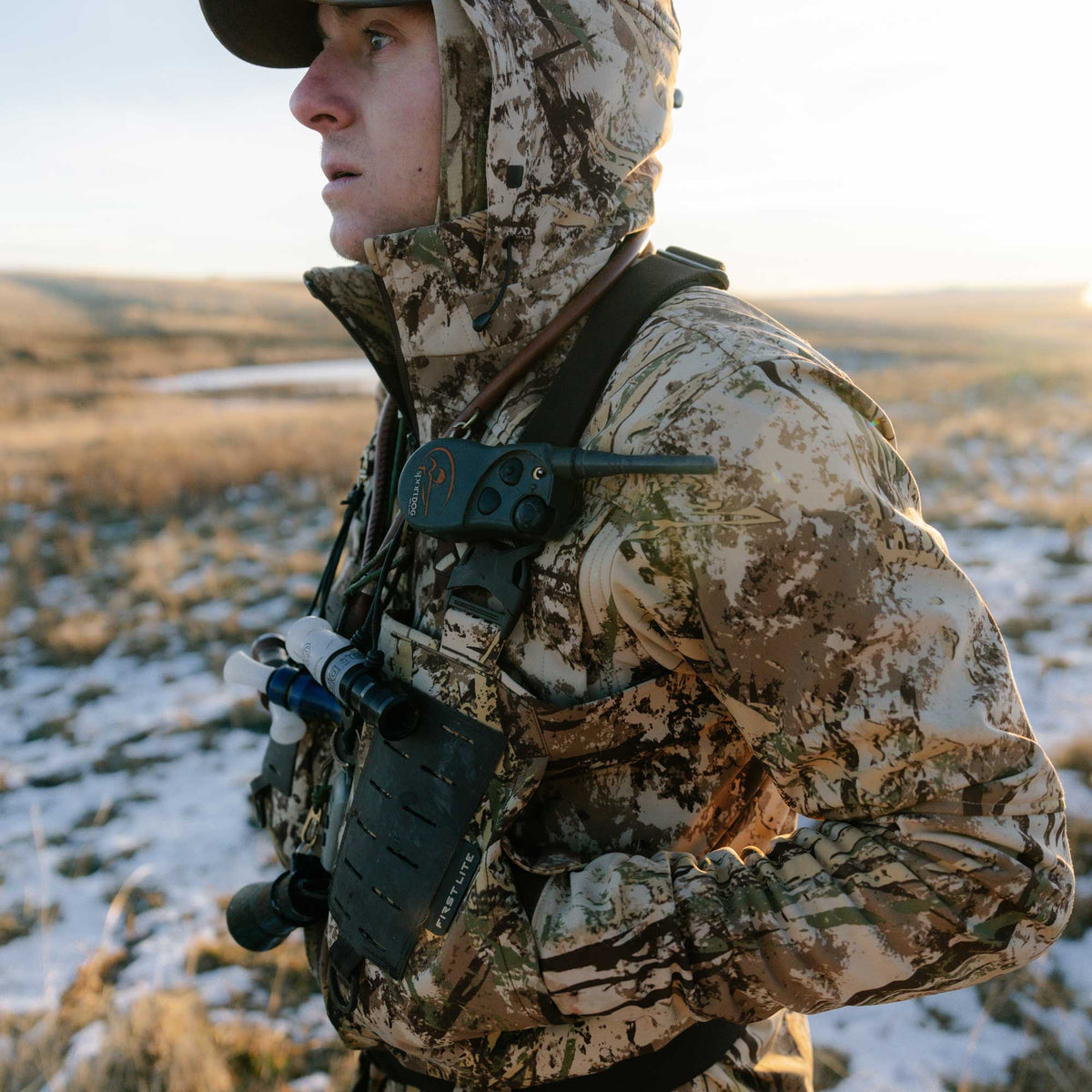 Hooded First Lite Fusion camouflage hunting jacket worn by a man with attached SportDOG device and calls, in a snowy field.