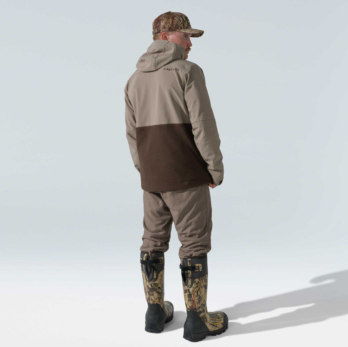 A man wears a two-tone tan and dark brown hooded jacket with 'FIRST LITE' logo, brown pants, a camouflage cap, and tall camouflage hunting boots, viewed from behind.