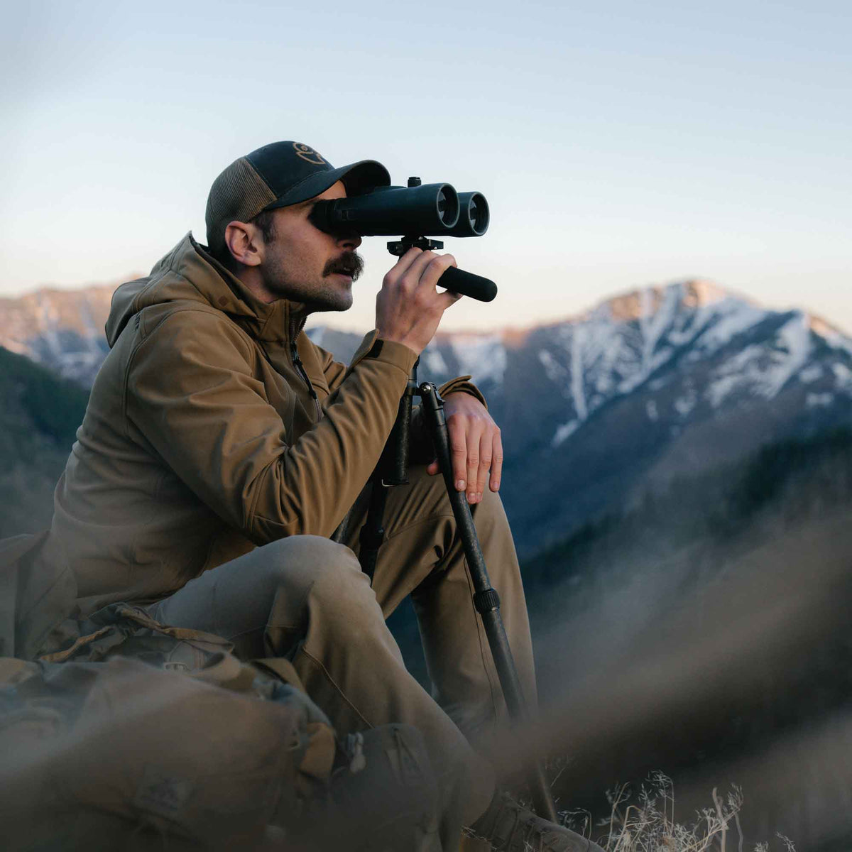A man uses large, dark binoculars mounted on a monopod to view a distant mountain range.