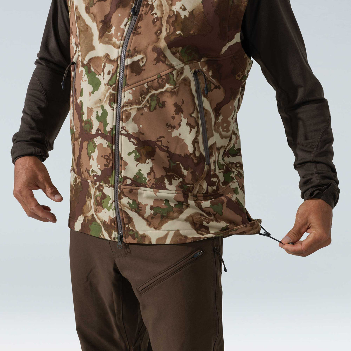 A person wears a multi-color camouflage vest with a full-zip front and chest pocket, adjusting the hem drawstring.