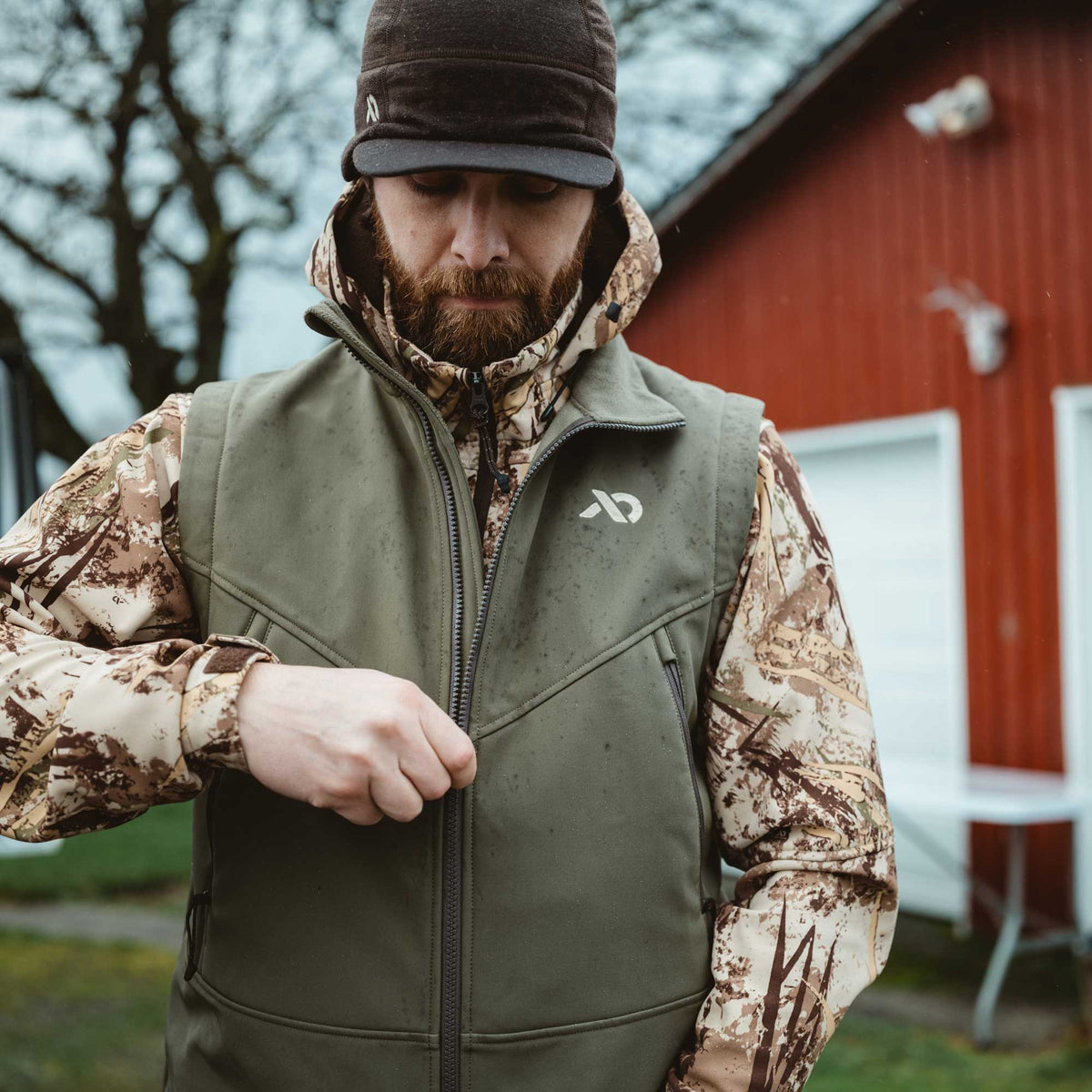 Man zipping up an olive green vest featuring a chest logo and zippered pockets, with water spots on the fabric.