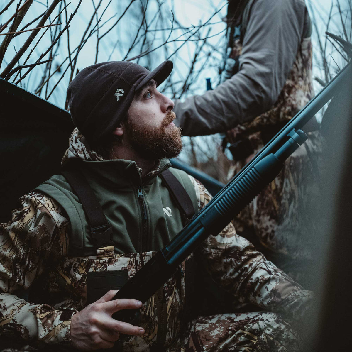 Man in a dark brown beanie with a logo, olive green zippered hunting jacket, and camouflage outfit holds a black shotgun outdoors.