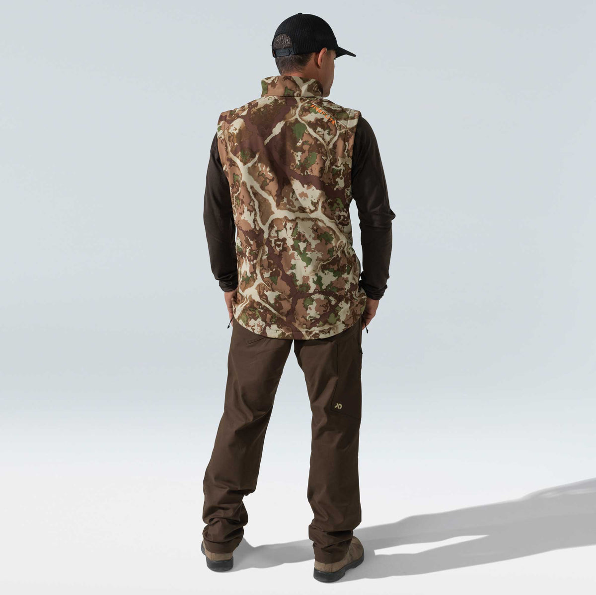 Man seen from the back wearing a First Lite camouflage vest, brown long-sleeve shirt, and brown pants.