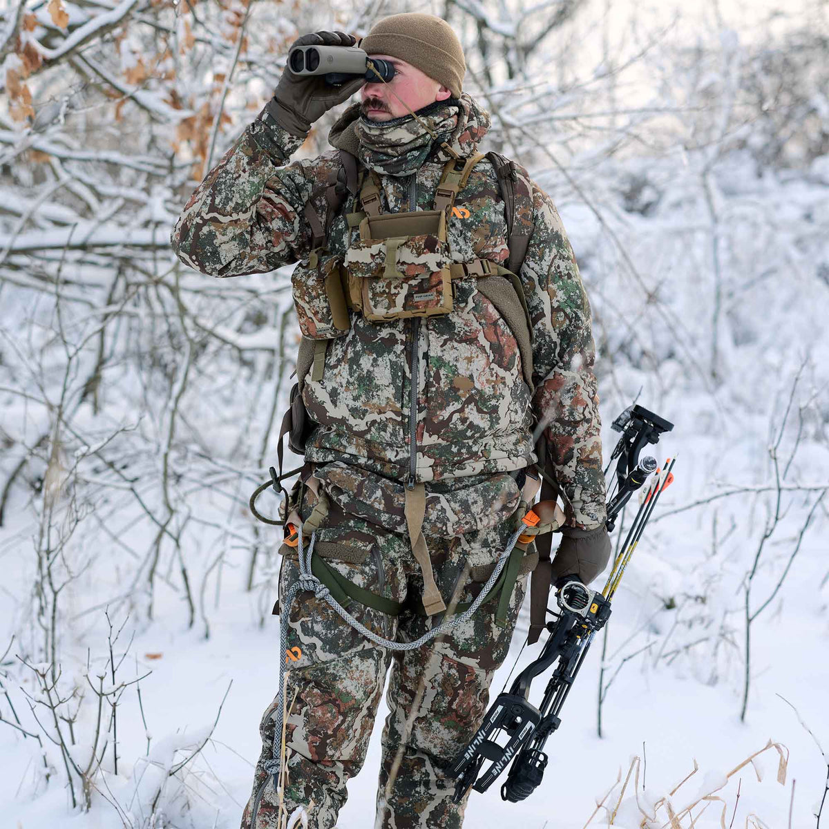 Man in multi-color camouflage hunting suit, beanie, gloves, chest rig, and harness, looking through binoculars in a snowy forest while holding a bow and arrows.