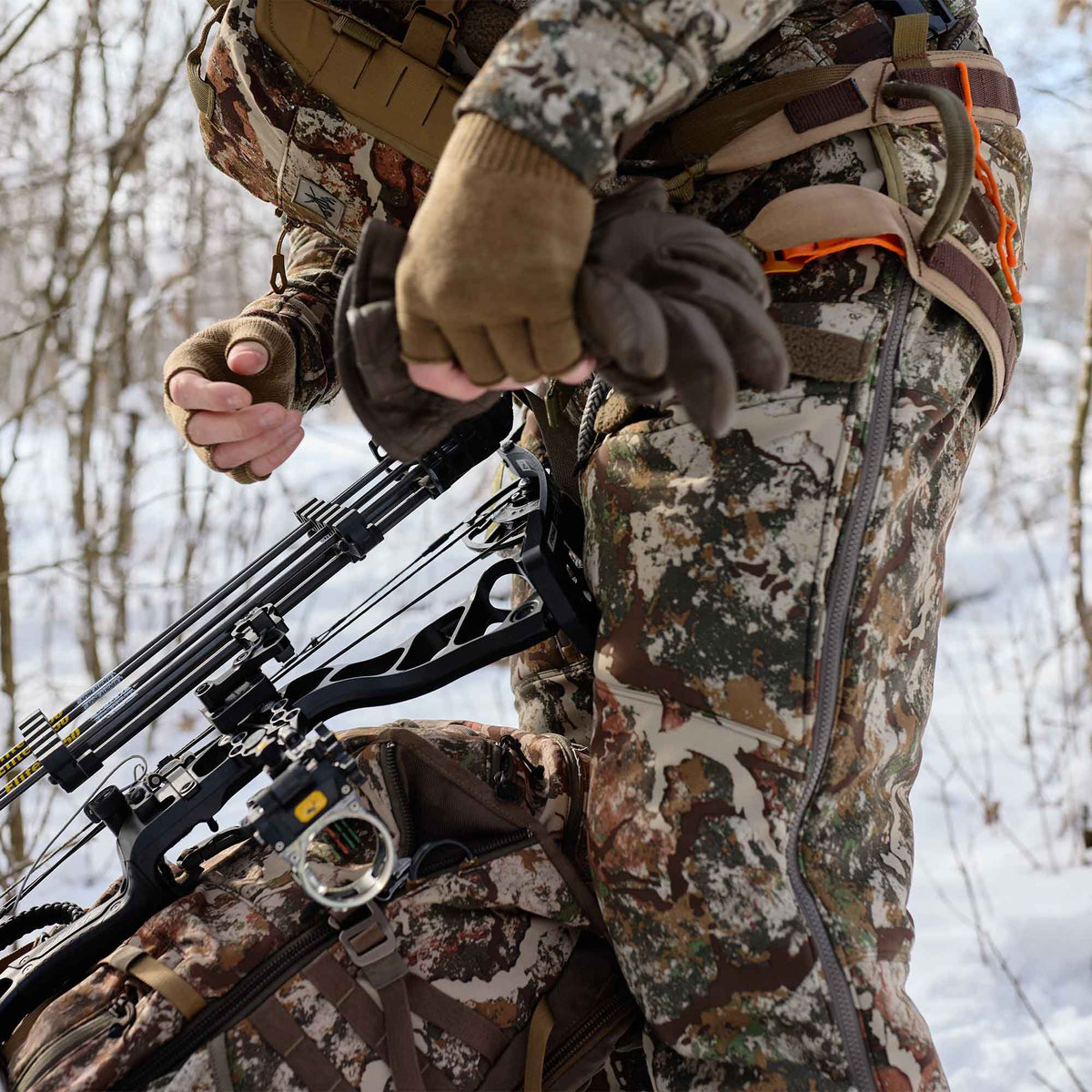 Hunter in winter camouflage hunting apparel and tan fingerless gloves, wearing a harness with orange accents, carrying a black compound bow with arrows and a matching camouflage backpack.