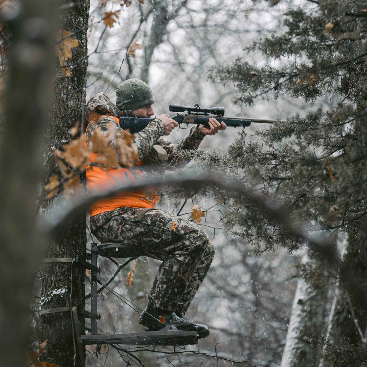 Hunter in a camouflage suit and orange vest aims a black and bronze rifle with a scope from a tree stand.