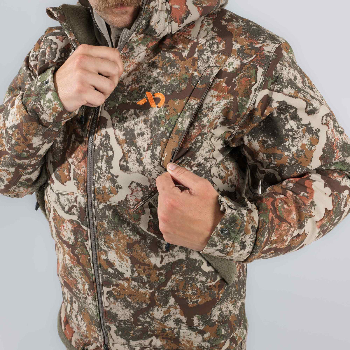 A person in a brown, green, and tan camouflage hooded jacket with an orange logo adjusts the main zipper and a chest pocket zipper.