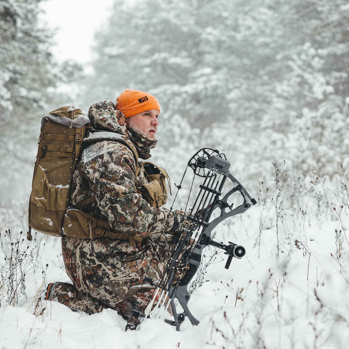 A hunter wearing camouflage winter gear and an orange beanie kneels in snow, holding a dark gray compound bow with a tan backpack.