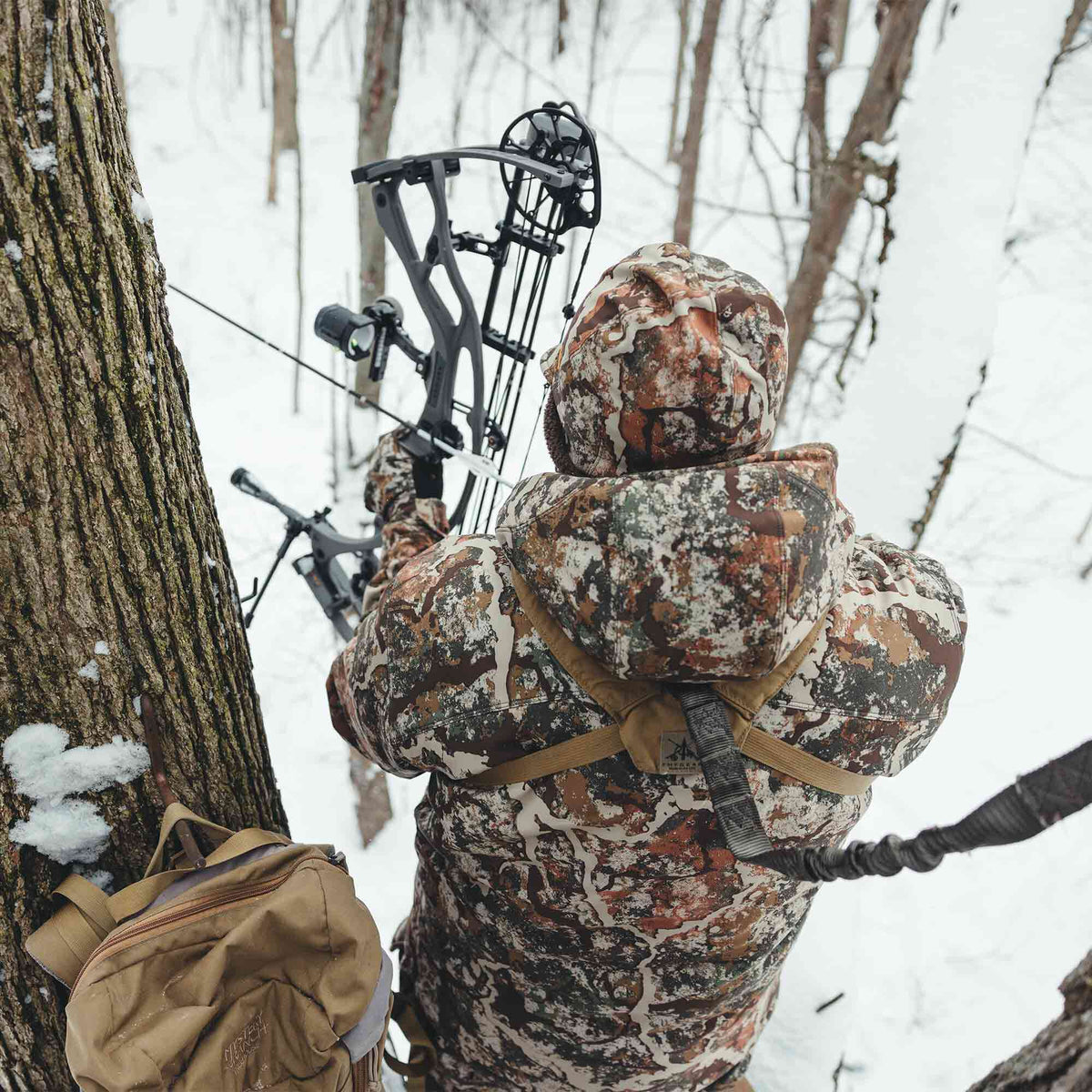 A hunter in a winter camouflage jacket and hood aims a black compound bow in a snowy forest, with a tan backpack hanging on a tree.