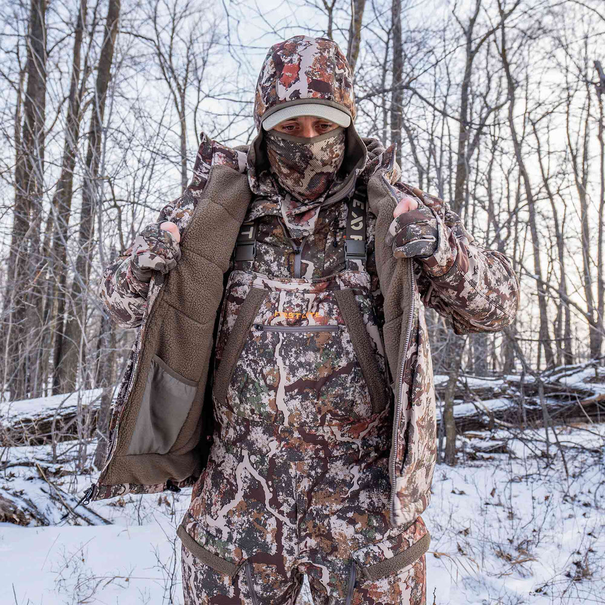 Earthy camouflage hunting suit featuring an open jacket with fleece lining, bibs with 'FIRST LITE' logo, a balaclava, and a hat, worn in a snowy forest.