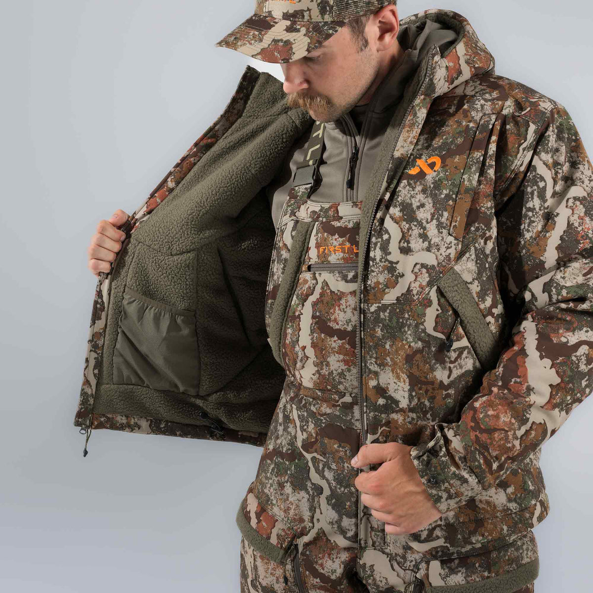 A man wears a brown, green, and tan camouflage hunting jacket with an orange logo, opened to reveal its thick olive green sherpa fleece lining and an internal mesh pocket.