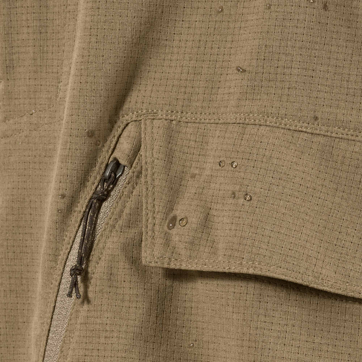 Khaki ripstop garment fabric showing a zipper, a flap pocket, and water droplets beading on the surface.