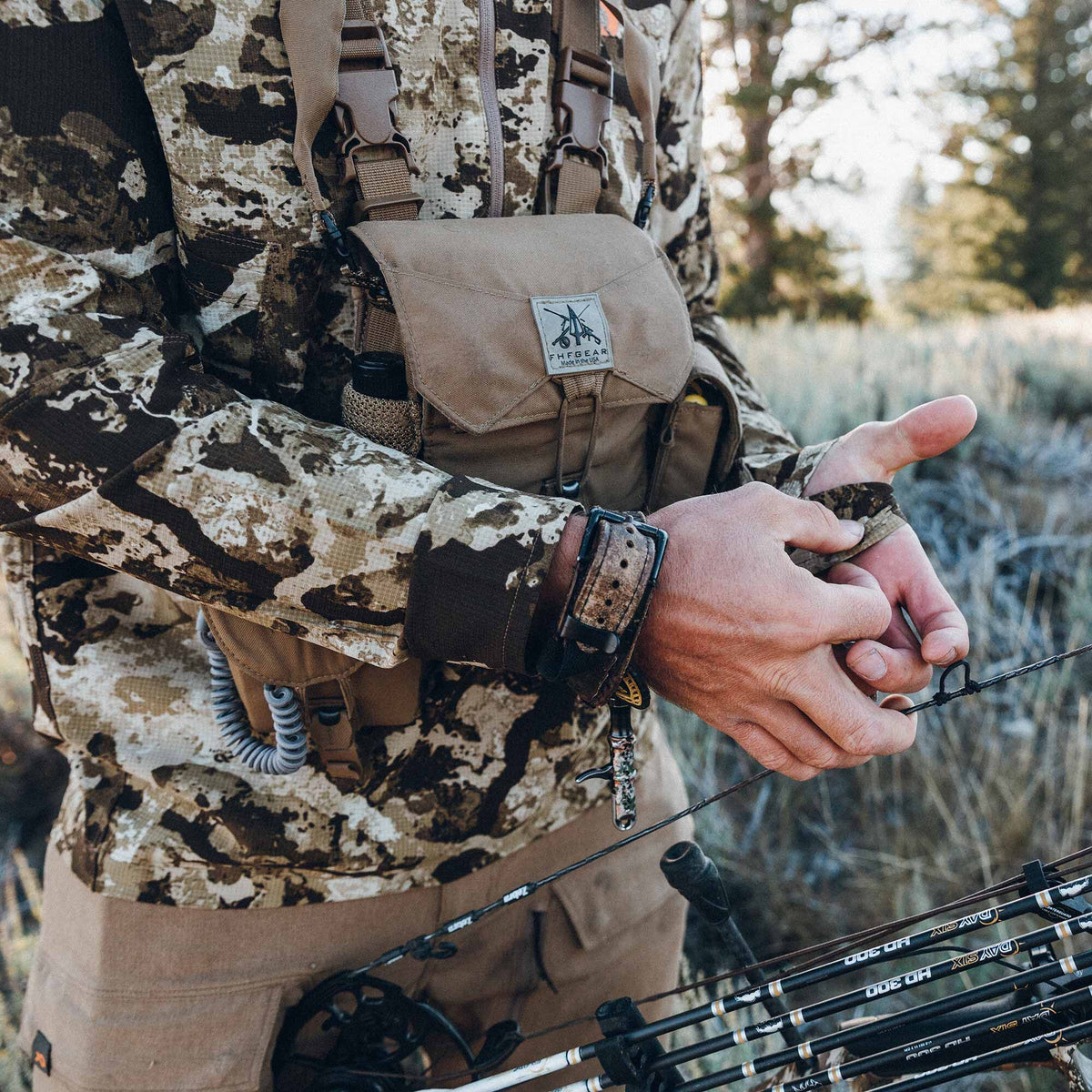 Tan FHFGEAR binocular chest pack with a prominent logo patch, worn by a person in a camo jacket holding a bowstring.