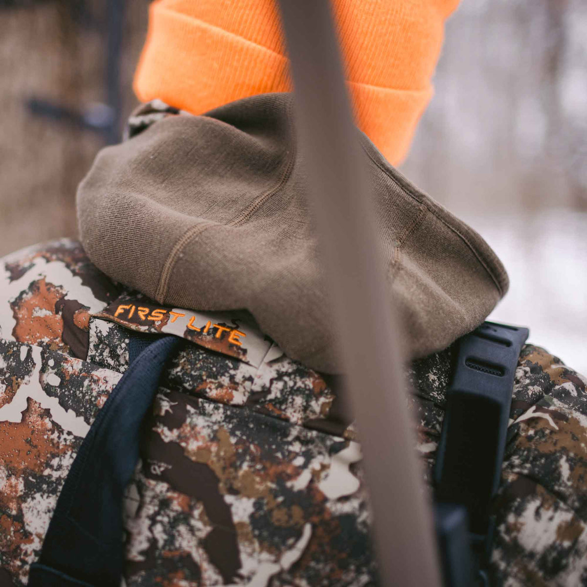 Man shown from behind wearing a camo jacket with FIRST LITE branding and an orange beanie.