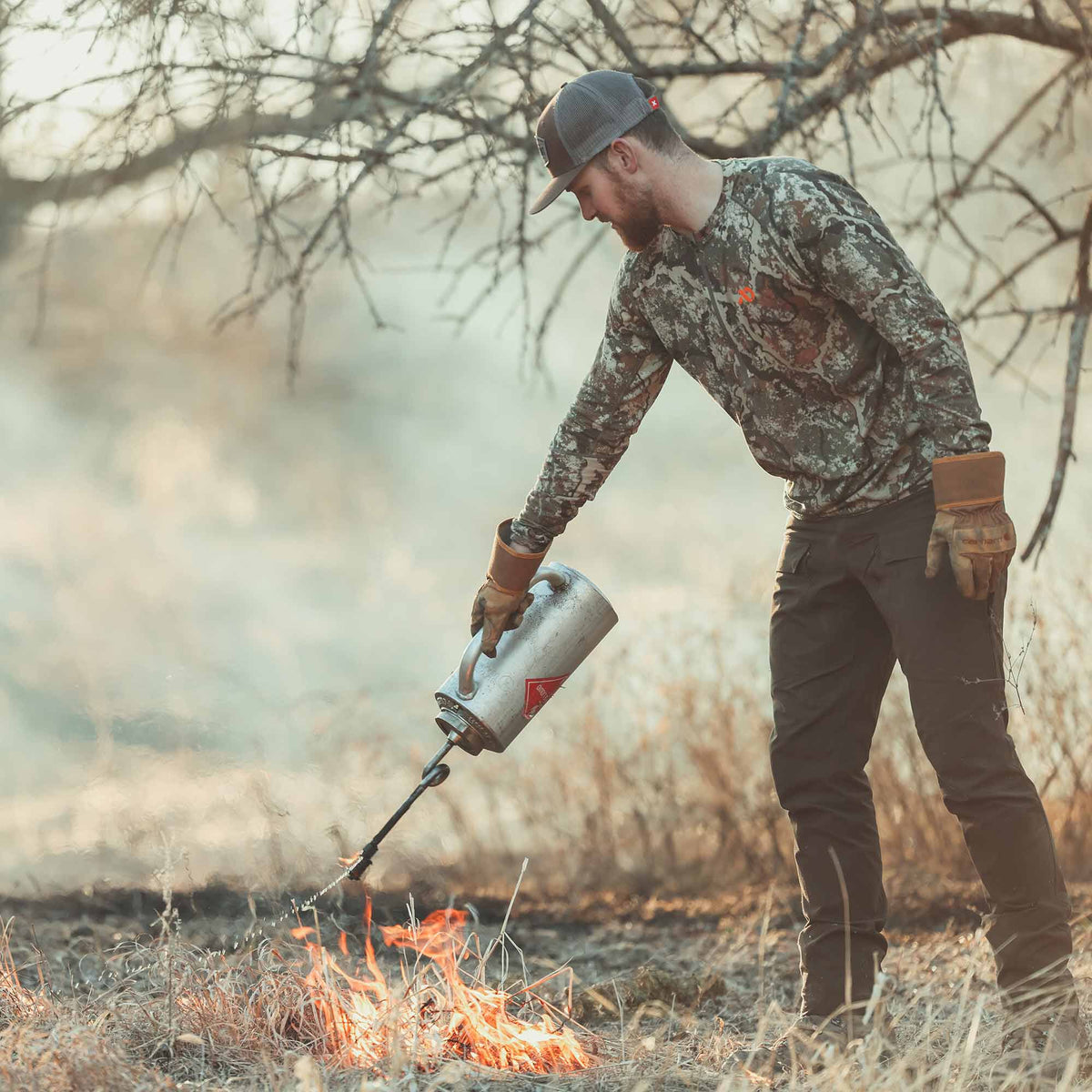 Silver drip torch with a red hazard label and long spout igniting dry grass.