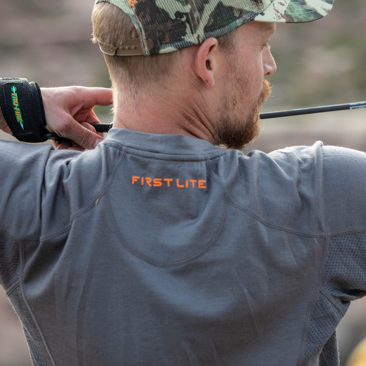 Dark gray First Lite long-sleeve shirt featuring an orange logo on the back and mesh side panels, worn by a man aiming a bow.