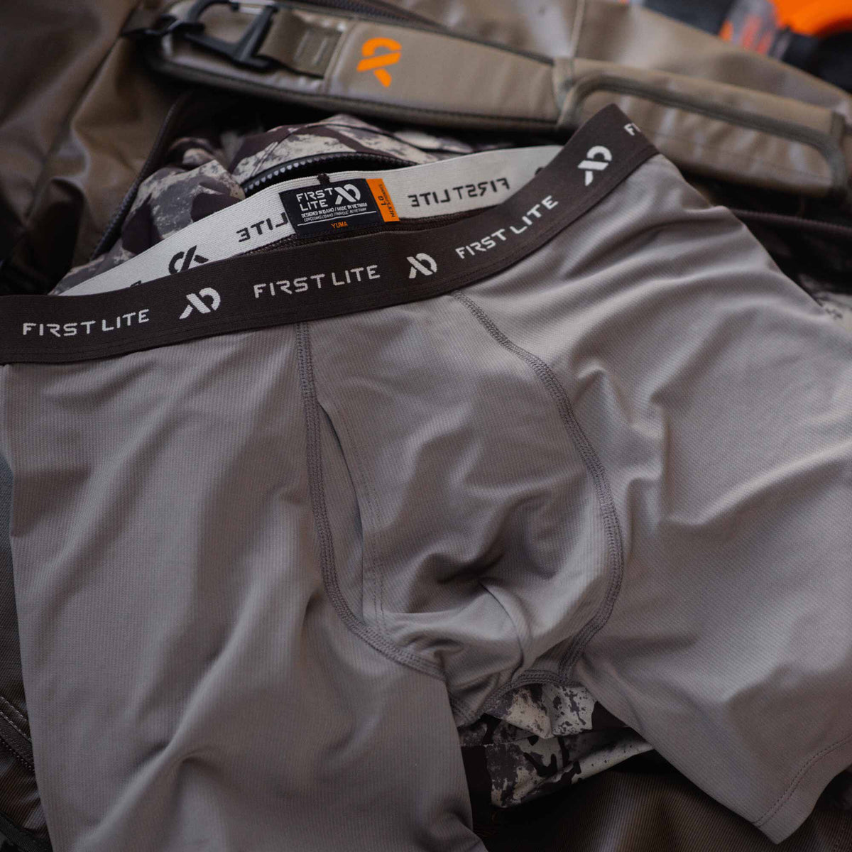 A pair of gray First Lite men's boxer briefs with a dark brown branded waistband and ribbed fabric texture.