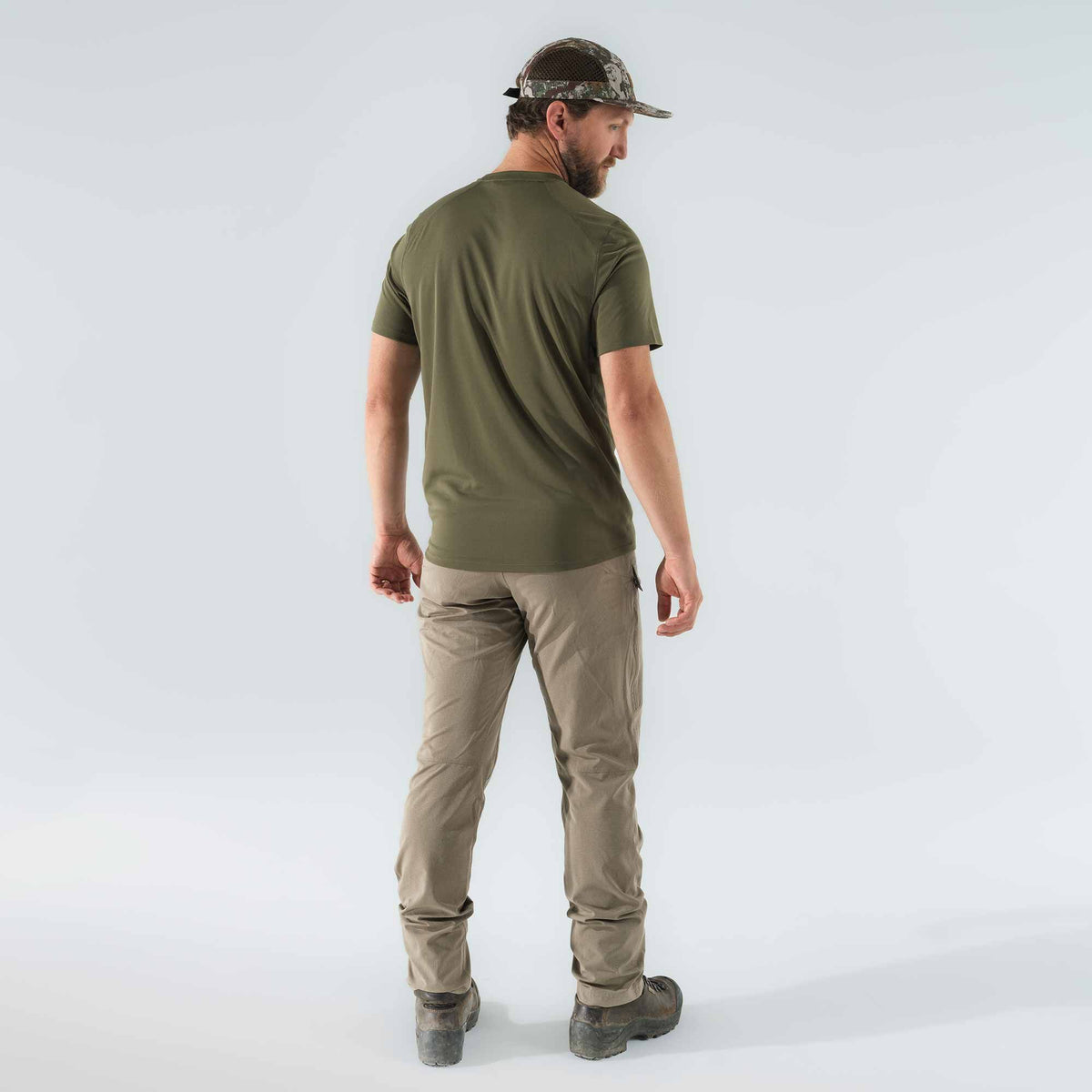 A man from behind wears an olive green t-shirt, tan cargo pants, and a camouflage mesh cap, looking over his right shoulder.