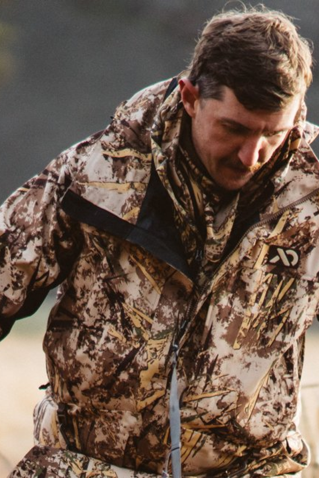 Man in brown camo jacket with black shoulder patch and AD logo on chest.