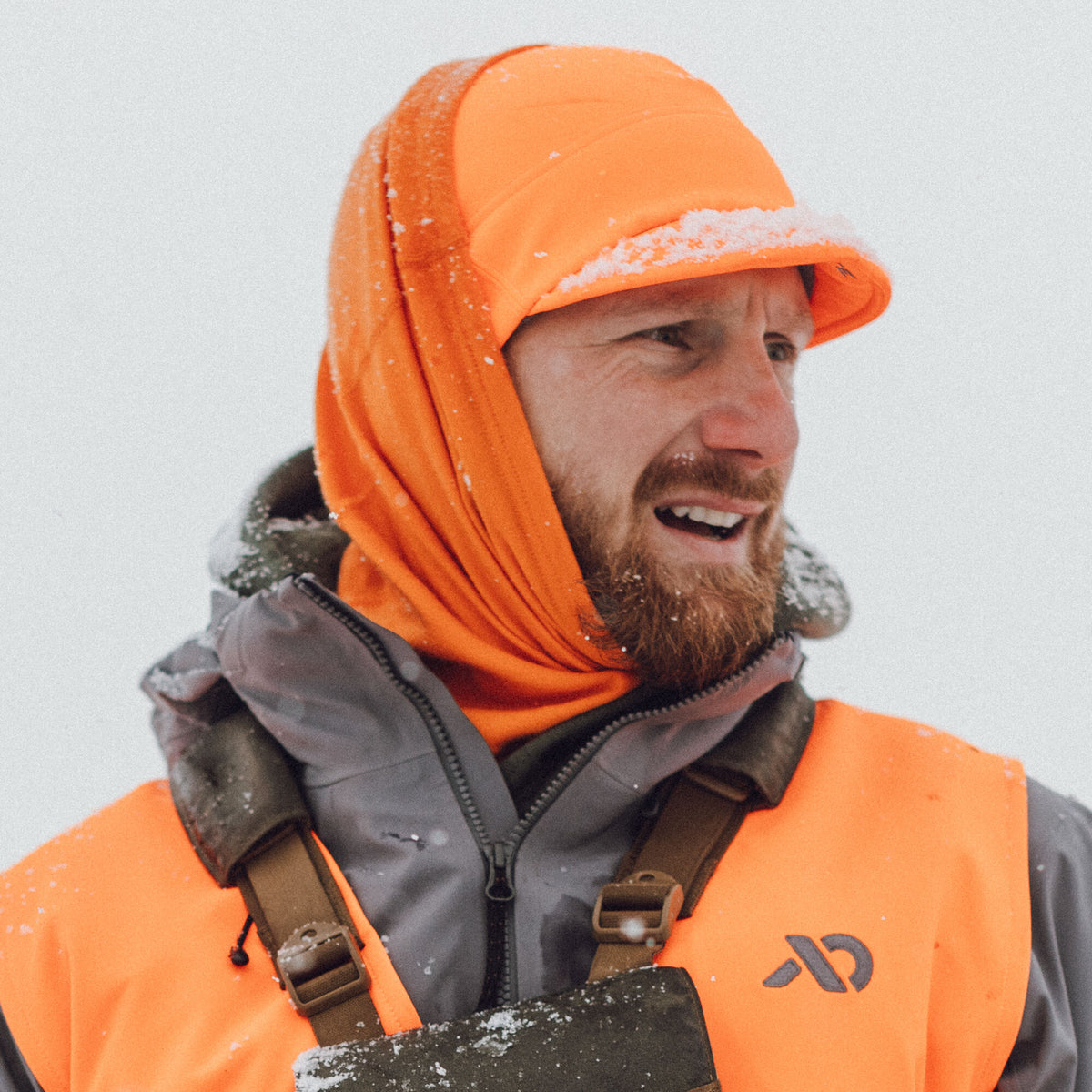 Close-up of a man wearing a snow-dusted blaze orange baseball cap, matching balaclava, and an orange hunting vest with a grey logo.