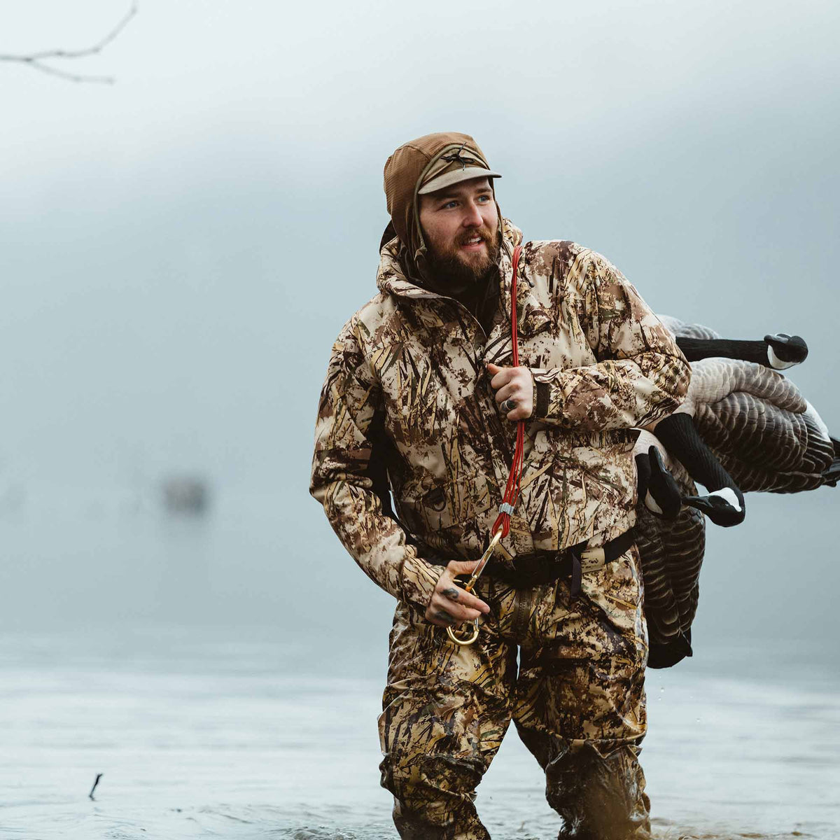 Brown, tan, and green marsh camouflage hunting jacket and pants worn by a man carrying goose decoys in water.