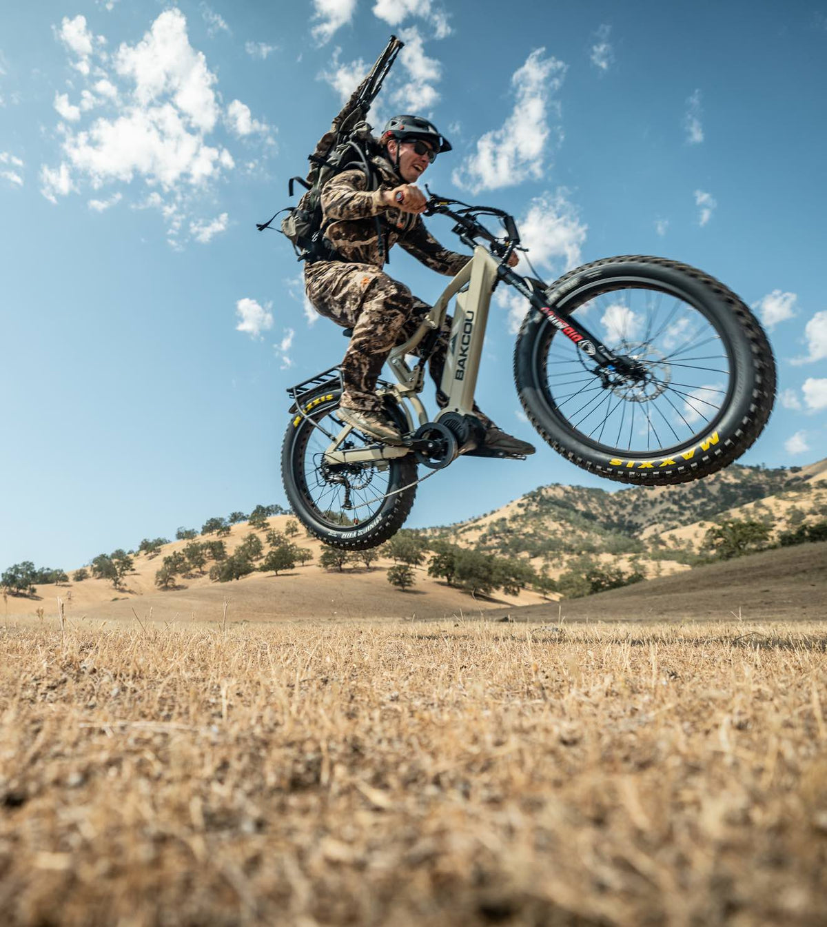 Hunter jumps tan Bakcou fat-tire e-bike with rifle in dry field.