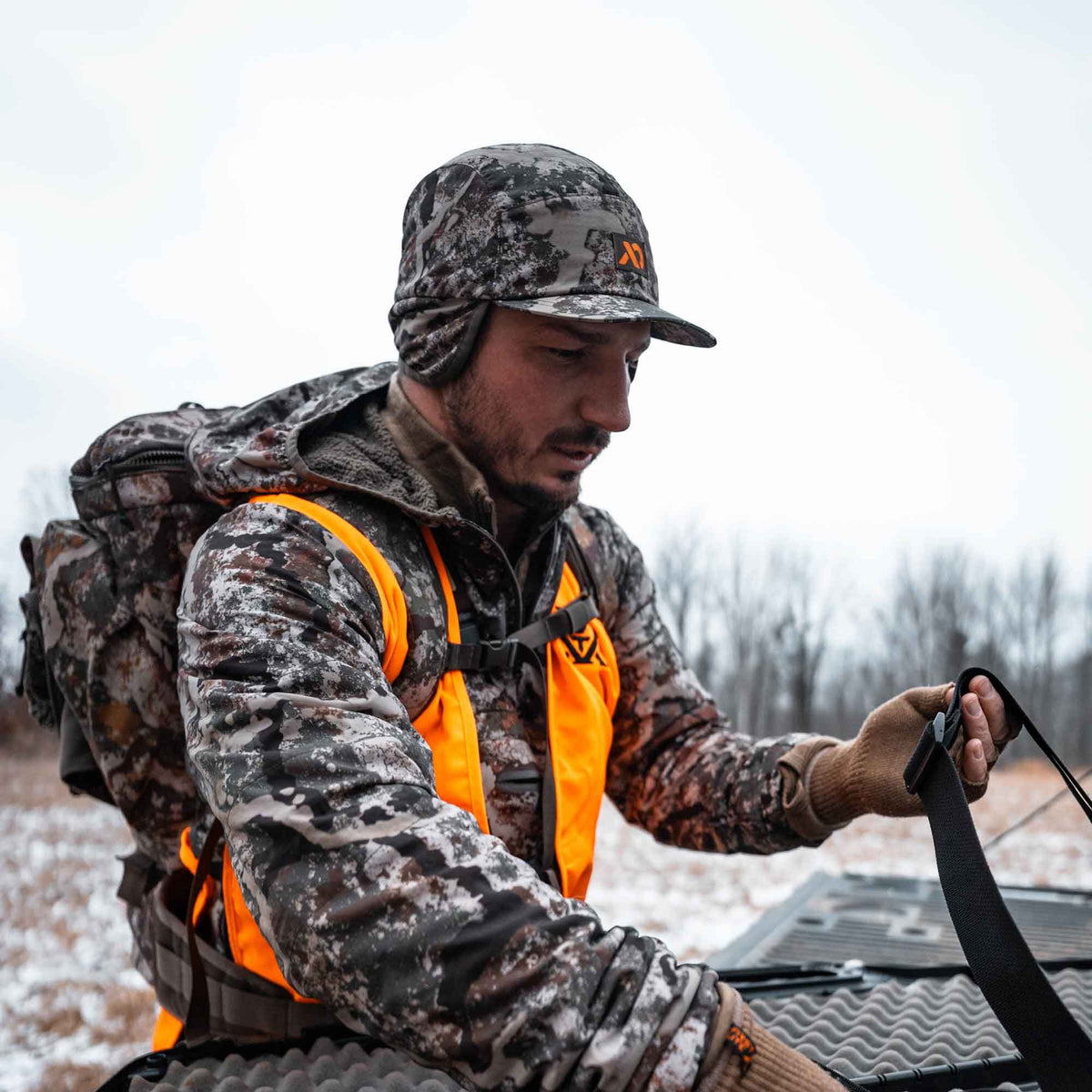 Camouflage hunting cap with ear flaps and orange logo worn by a man also in a bright orange safety vest and camouflage jacket.