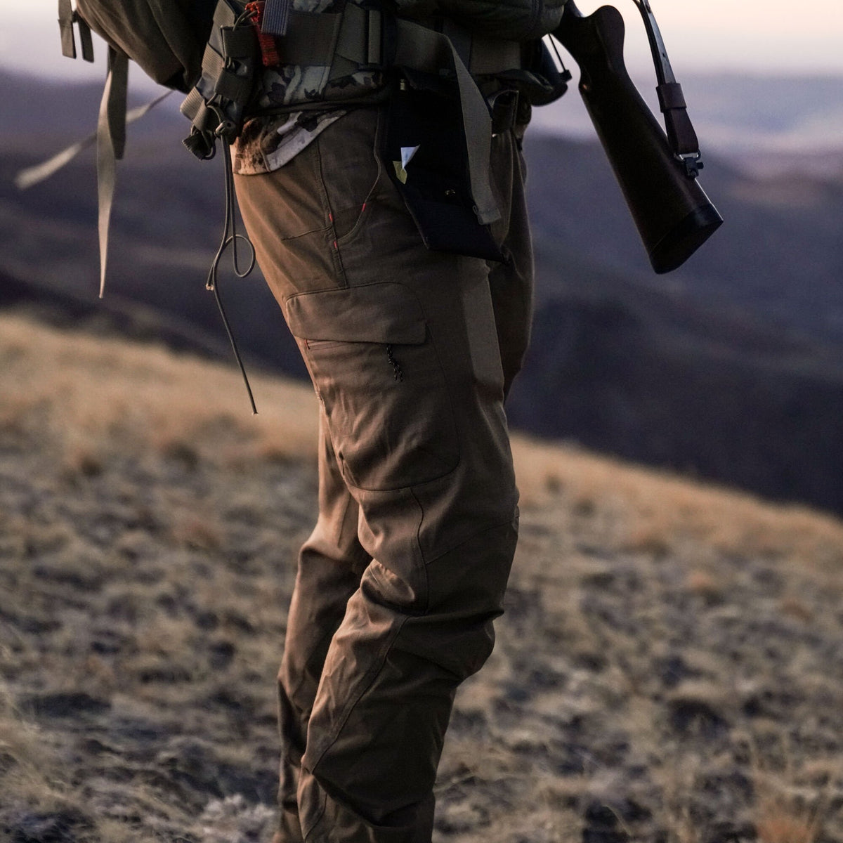 Brown outdoor pants with zippered cargo pockets and red stitching, worn by a person carrying a rifle.