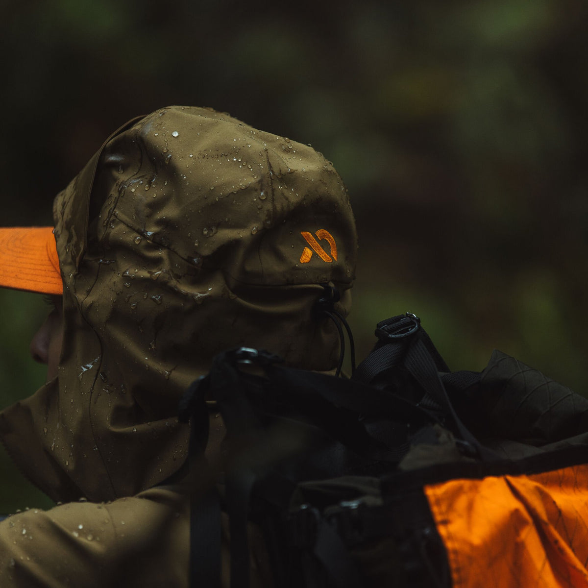 Olive green water-repellent hood with an orange logo and drawstring, worn over an orange cap.