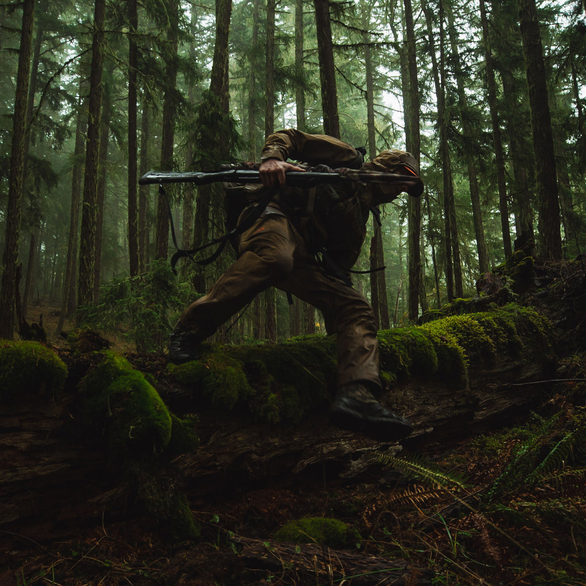 A person in brown outdoor gear with a dark backpack and rifle steps over a mossy log in a dense forest.