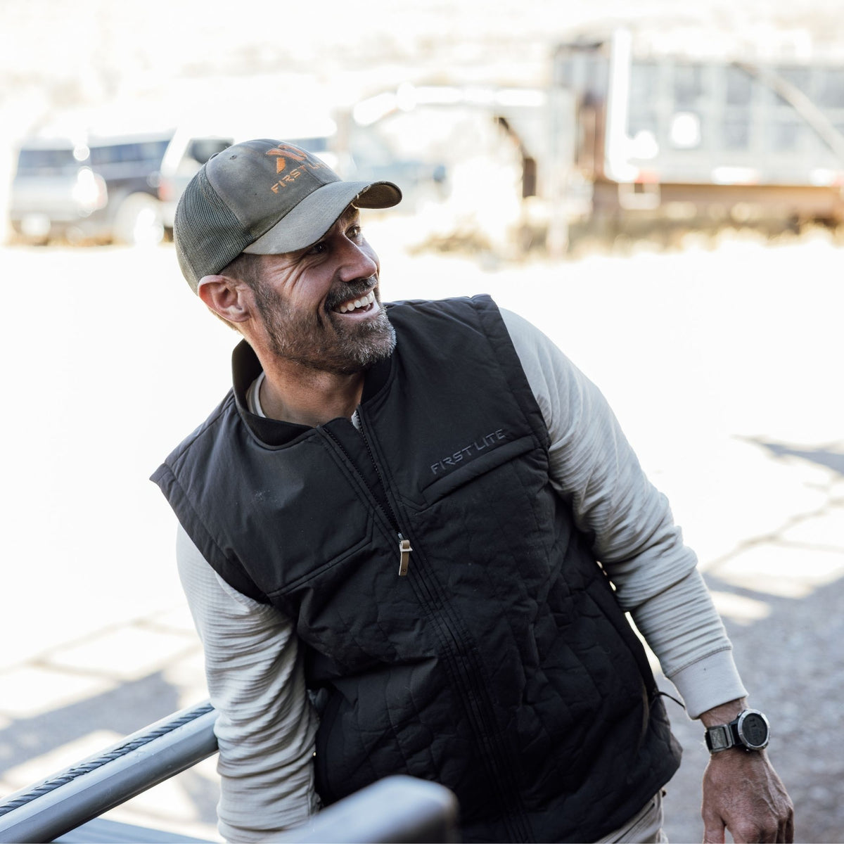 Man wearing a black outpost vest over a light gray shirt and a gray cap with FIRST LITE branding.