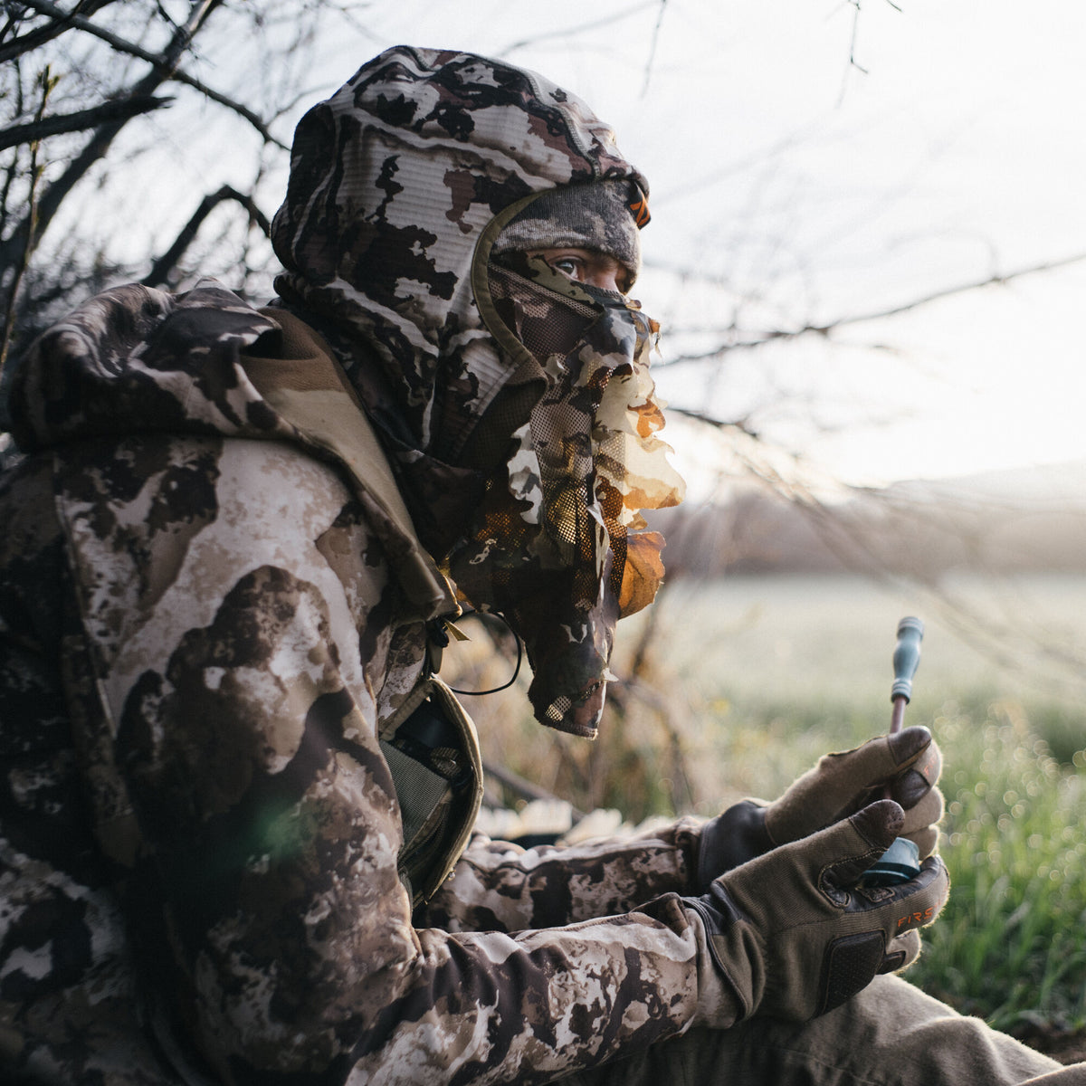 A person in brown and white multi-camouflage hunting apparel with a hooded top and leafy mesh facemask holds a gray game call outdoors.