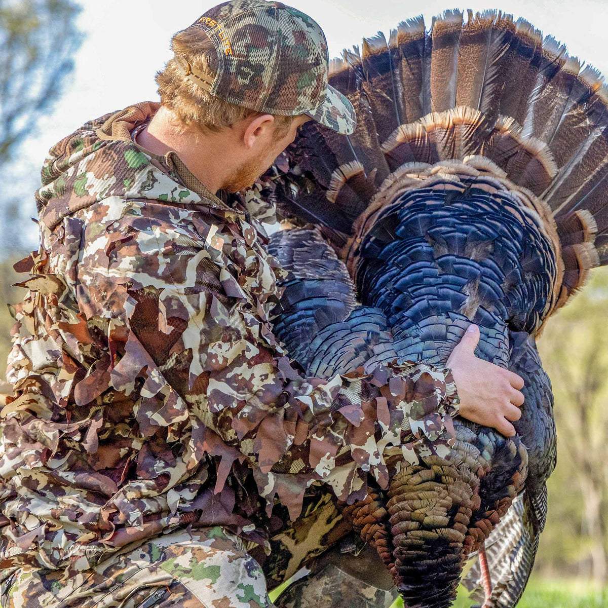 Person in a brown, green, and tan leafy camouflage jacket and mesh-back hat holding a large wild turkey with iridescent blue body feathers and a fanned brown tail.