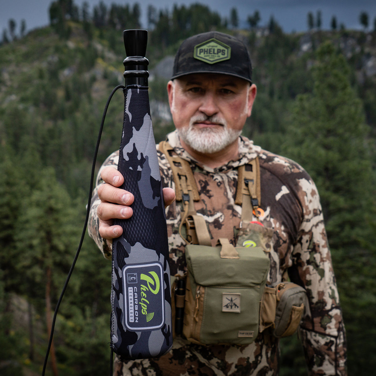 A hunter holds a black and grey camouflage Phelps Carbon Technology game call with a black mouthpiece.