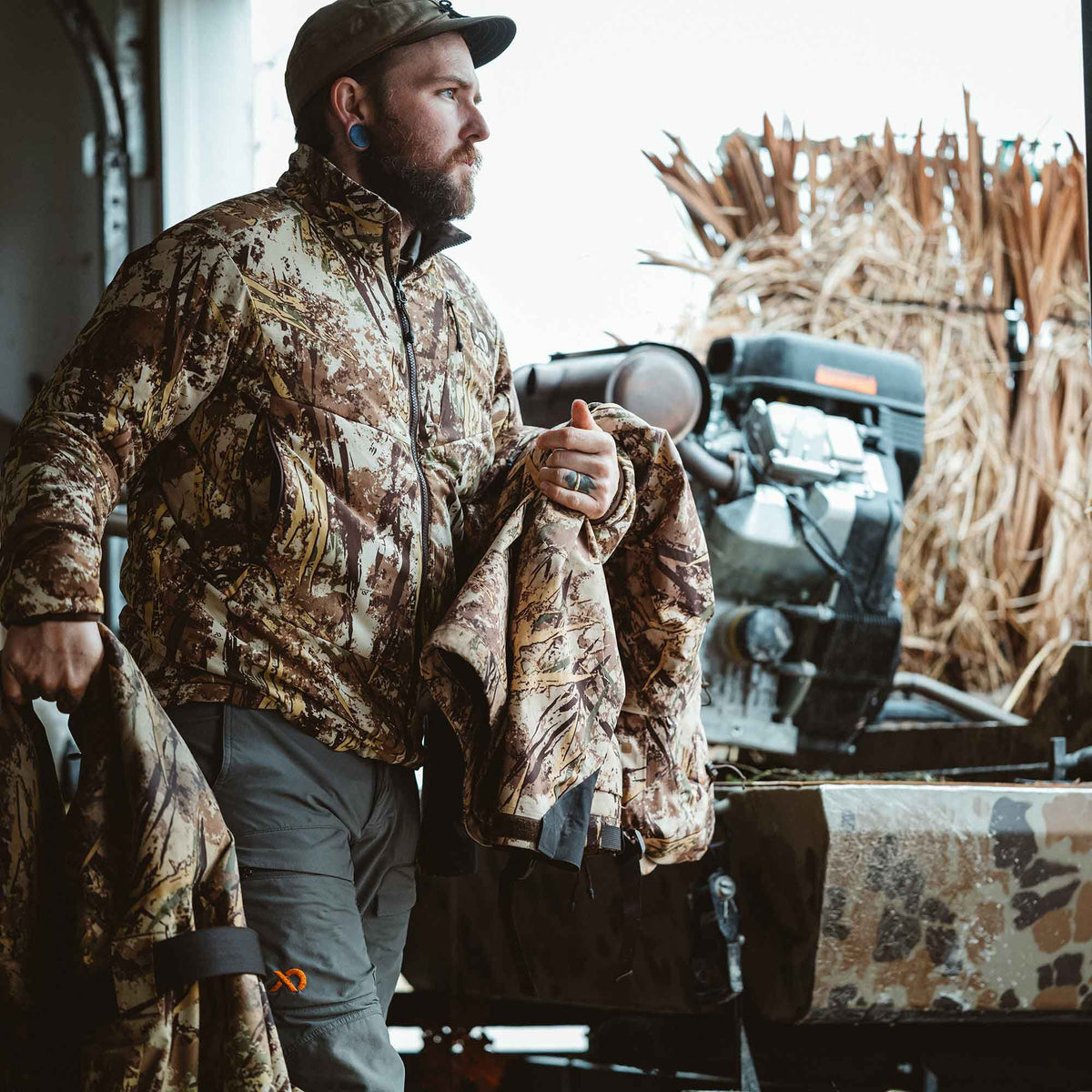 Man wearing a brown, tan, and black camouflage zip-up jacket with a stand collar, holding another matching apparel item.