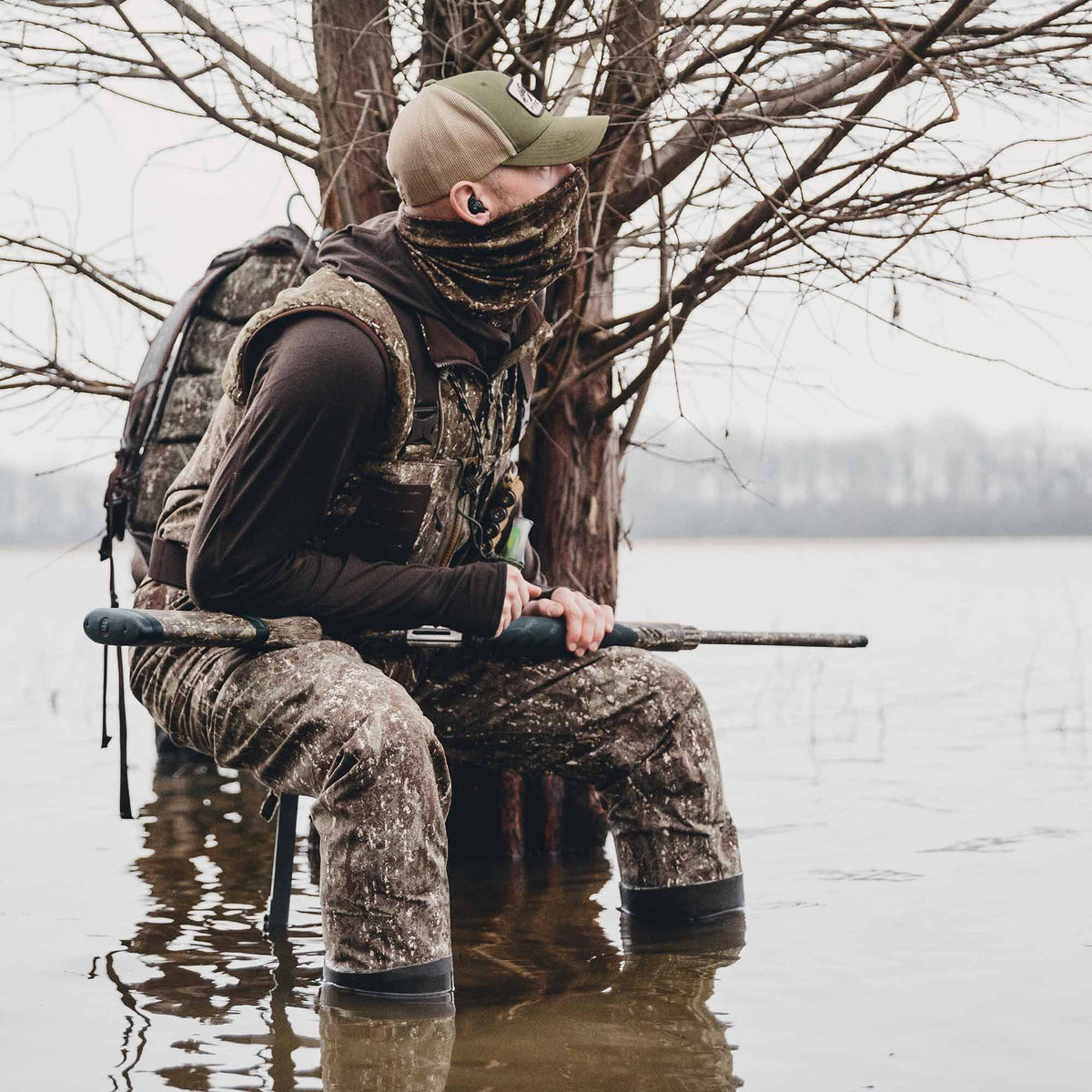 Hunter in full camouflage waders, vest, and neck gaiter sits in water, holding a shotgun and wearing earbuds.