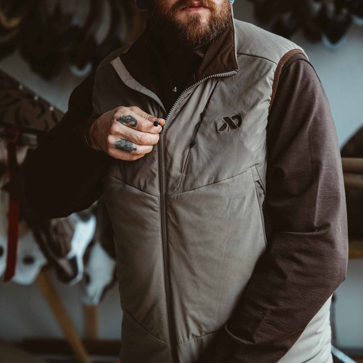 A person zips up a Walnut padded vest featuring a full-length zipper, dark brown collar and shoulder trim, and an embroidered chest logo.