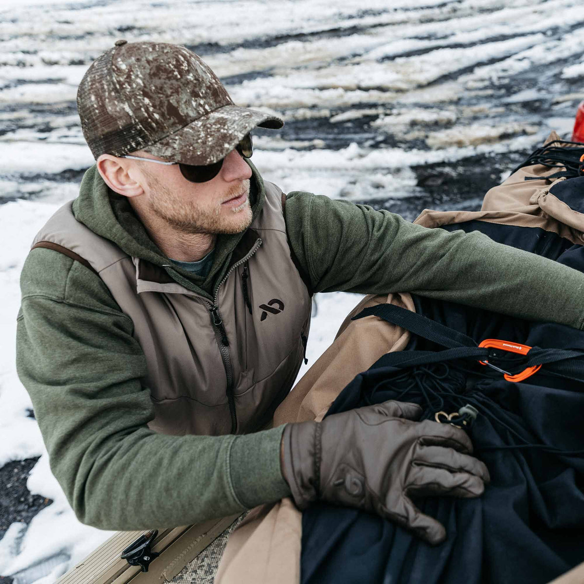 Tan outdoor vest with a black chest logo and dark zipper, layered over an olive green hoodie.