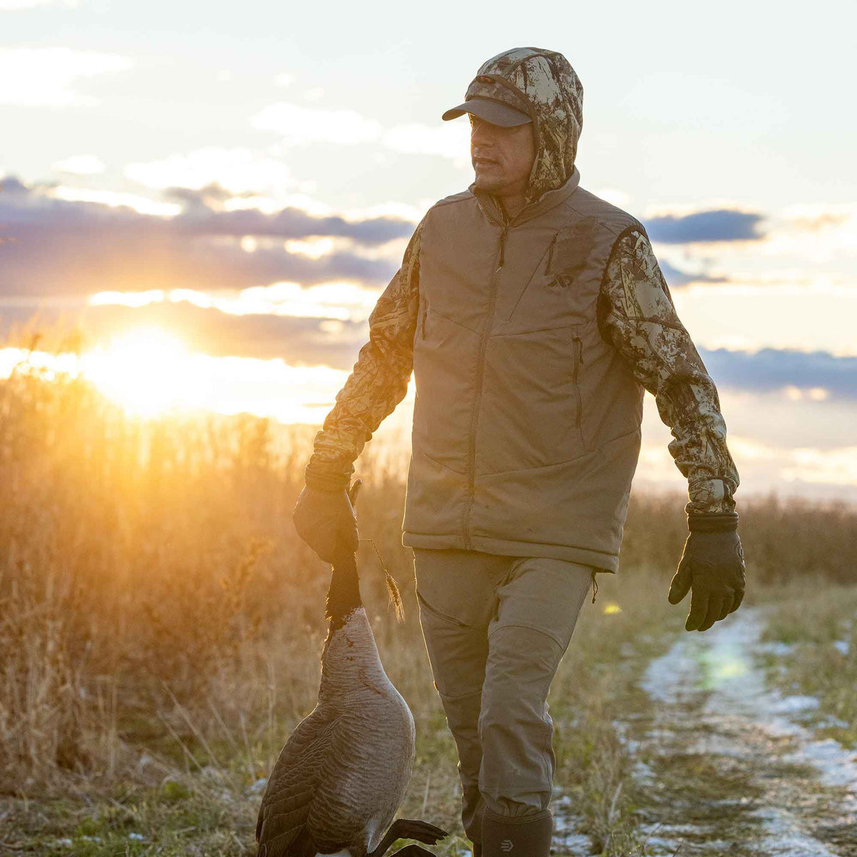 Man in a brown full-zip hunting vest over a camouflage shirt and cap carries a goose on a snowy path at sunset.