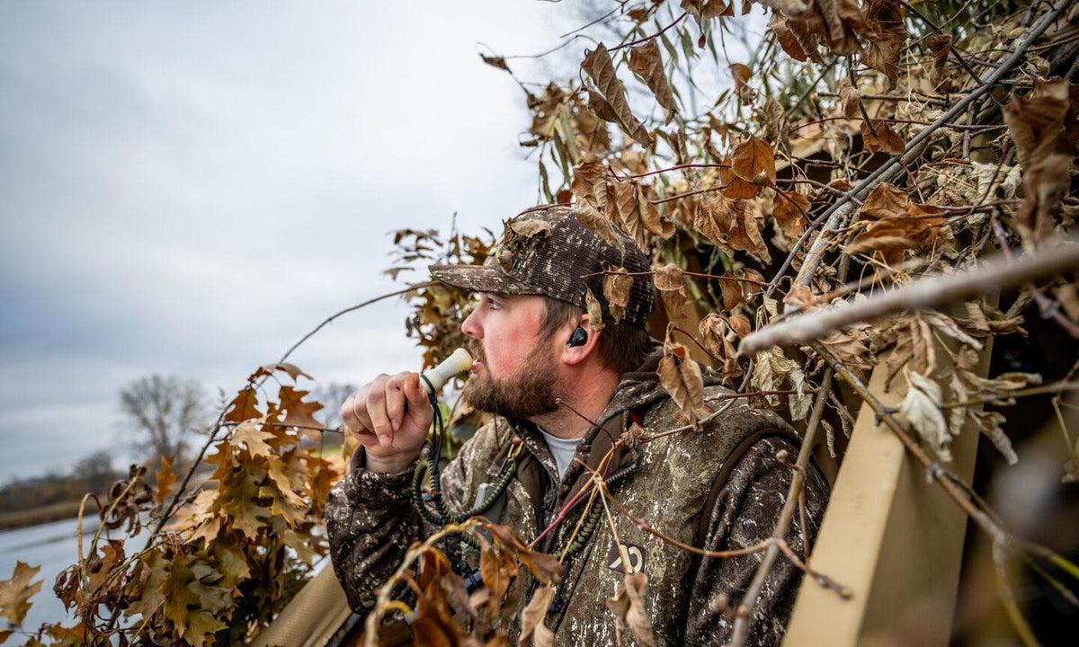 Camo hunter blowing off-white duck call, wearing dark ear protection earbuds.