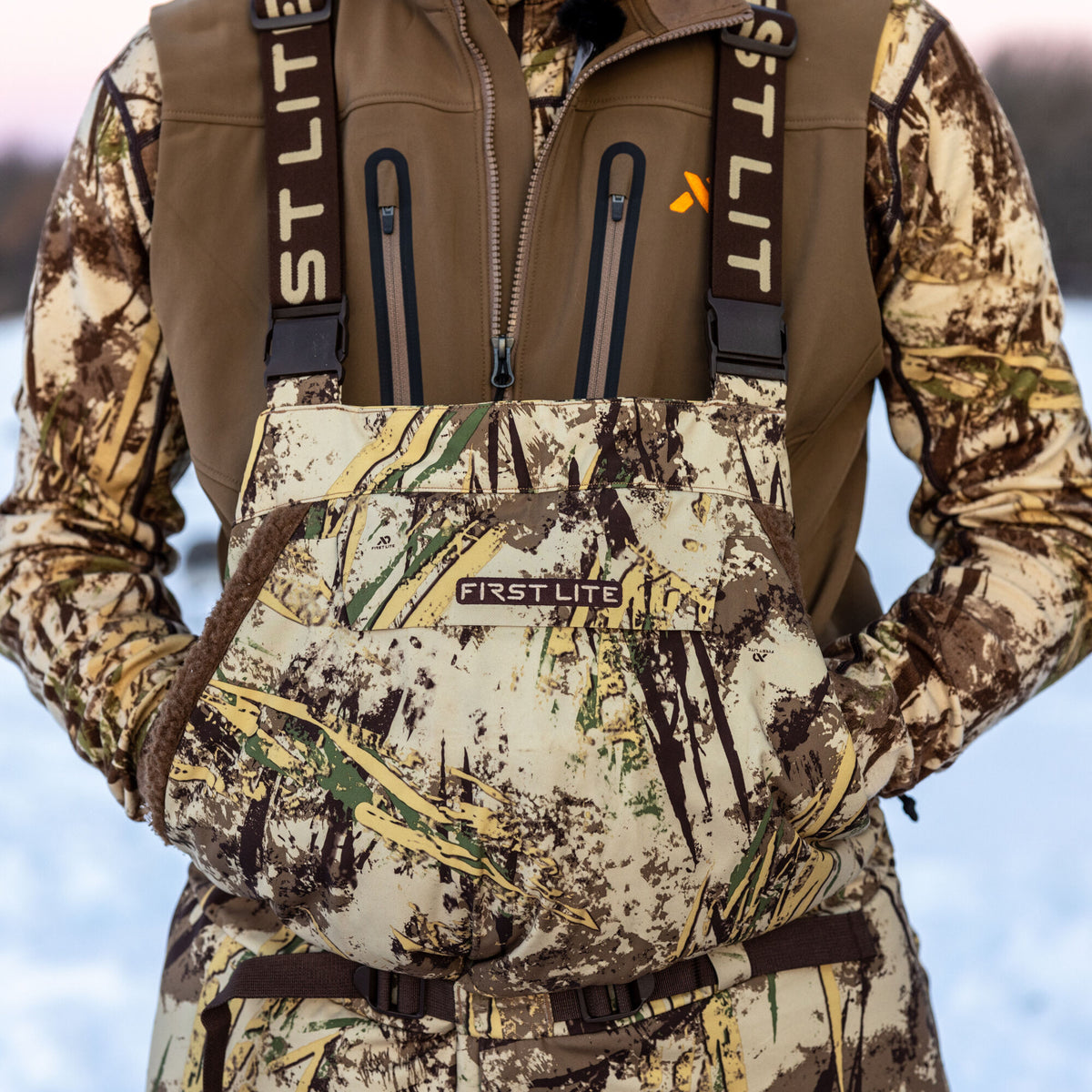 First Lite camouflage hunting bibs feature a large central handwarmer pocket with brown fuzzy trim and brown branded suspenders.