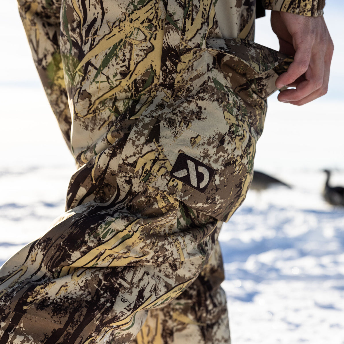 Close-up of camouflage hunting pants in muted earth tones with a visible logo and a hand reaching into a side pocket.