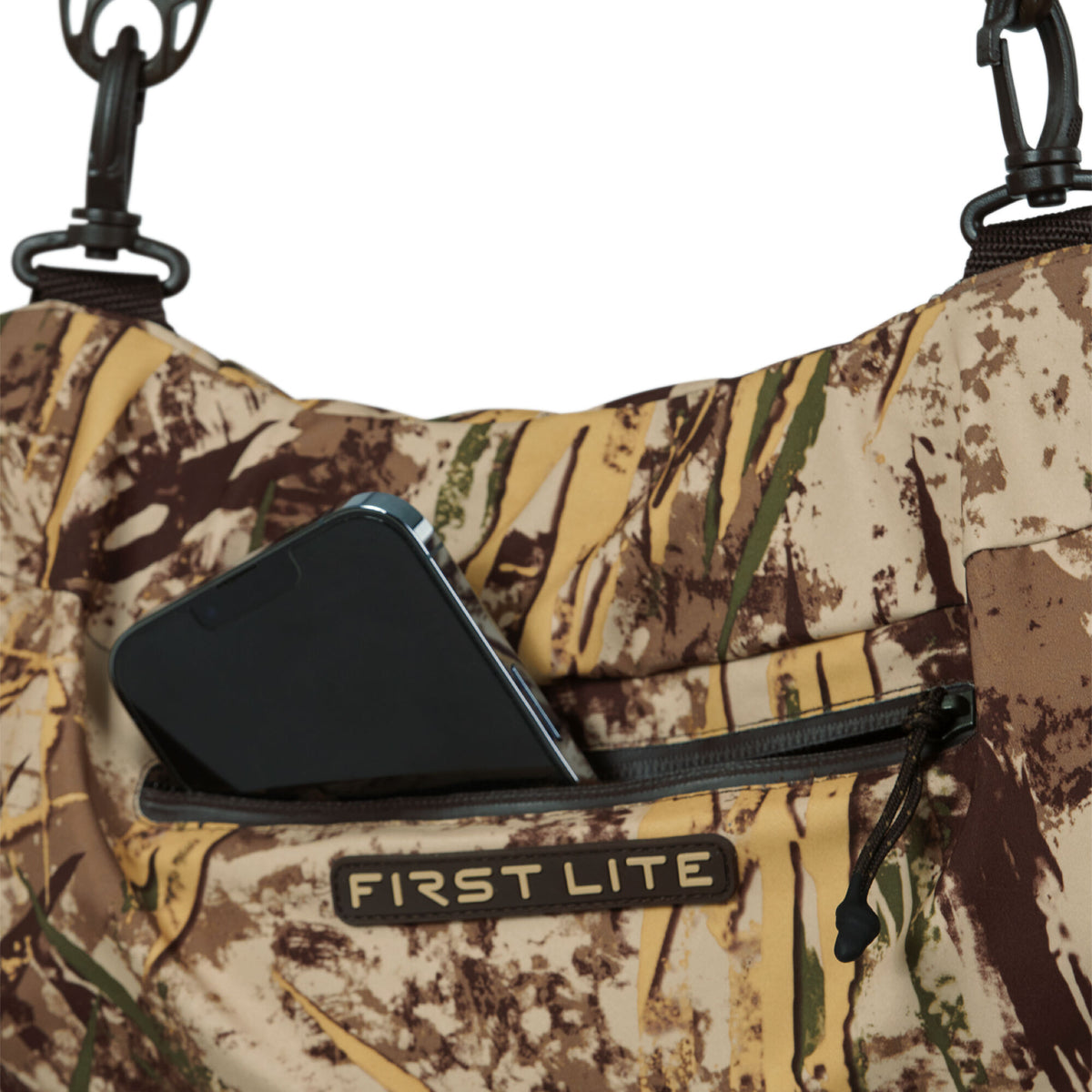 First Lite camouflage outdoor gear featuring a zippered pocket holding a smartphone, with earthy tones and black strap clasps visible.