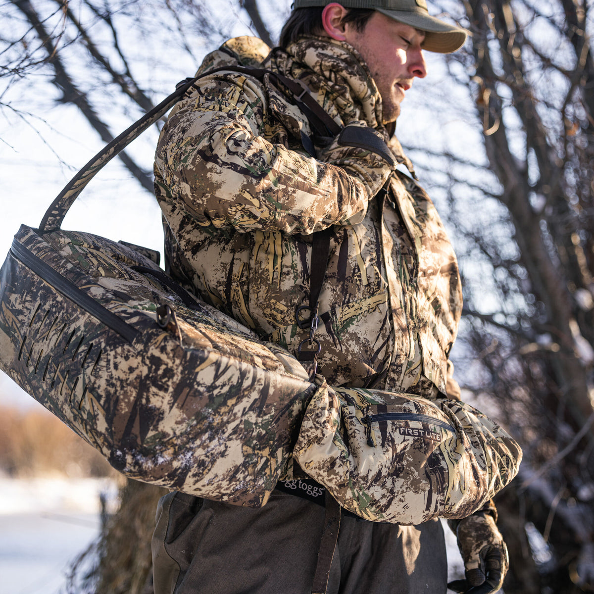 Brown and green camouflage duffel bag with perforated side panel and matching First Lite hand muff, carried by a person in a matching camouflage jacket.